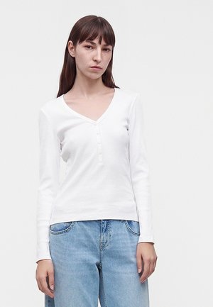 Ung person med glat brunt hår iført en hvid langærmet V-hals trøje og lyseblå jeans foran en ensfarvet baggrund.