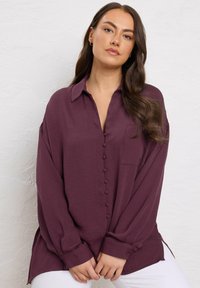 Donna con lunghi capelli castani che indossa una blusa viola buttonata larga e pantaloni bianchi, in piedi contro un muro bianco testurizzato.