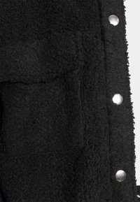 Svart fleece-tyg med en mjuk, texturerad yta. Har silverfärgade tryckknappar längs kanten. Inga distinkta mönster eller ytterligare dekorationer.