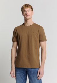 Bruin T-shirt met korte mouwen van zachte stof, met een ronde halslijn en een klein logo op de borst; gecombineerd met blauwe jeans.