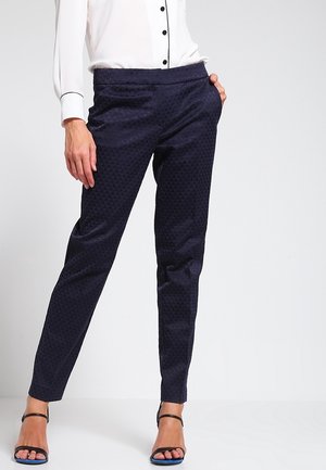 Femme portant un pantalon slim foncé à motifs, un chemisier blanc avec des boutons noirs et des escarpins noirs ouverts à l’avant, debout avec une main dans la poche.