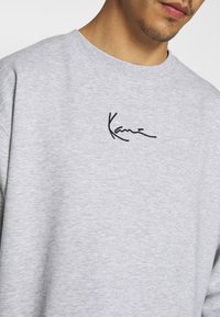 Grå sweatshirt fremstillet af blødt stof, med rund hals og et broderet sort signaturdetalje på brystet.