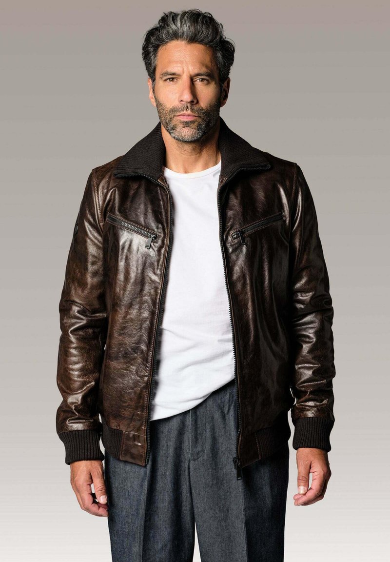 Veste en cuir marron avec fermeture éclair à l'avant, col haut et poignets côtelés. Comprend deux poches zippées et une finition lisse et texturée.