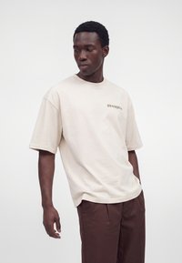 Homme portant un t-shirt beige ample avec le texte "DROPOUT" et un pantalon marron, regardant vers sa gauche sur un fond blanc uni.