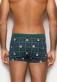 Boxer shorts a cuadros verdes con patrones de personajes de dibujos animados en blanco y rojo, que presentan un ajuste ceñido y una cinturilla elástica verde.