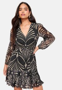 Rochie neagră cu mâneci lungi, cu decolteu în V adânc, decorată cu un model bej cu frunze. Material fluid din chiffon cu tiv ondulat și talie strânsă.