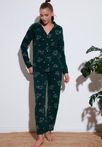 Femeie stând desculță îmbrăcată într-un costum de pijama verde închis cu nasturi, cu imprimeuri de ursuleți de pluș și stele, pe lângă un perete alb, lângă o plantă.