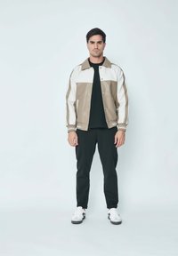 Veste en cuir kaki et blanc avec un col, dotée de boutons-pression et de poignets côtelés. Chemise noire en dessous et pantalon noir. Baskets blanches.