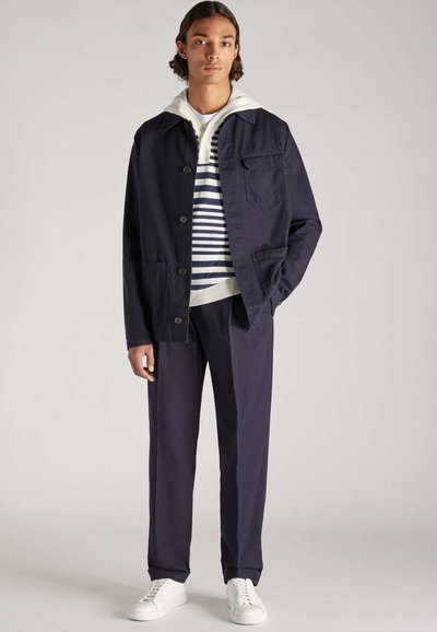 Marineblaues Jackett mit Knopfverschluss, einer Brusttasche, über einem gestreiften Pullover und grauem Hoodie getragen, kombiniert mit marineblauen Hosen und weißen Sneakers.