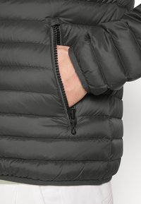 Handen i den dragkedjade fickan på en svart vadderad jacka med horisontell sömnad, bärs över vita byxor.