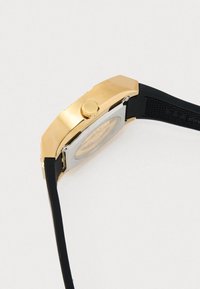 Relógio em tons de ouro e prata com caixa hexagonal, pulseira de borracha preta e botão de pressão lateral; possui uma pulseira texturizada e um mostrador refletivo.