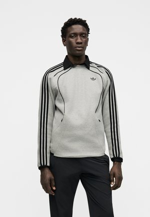 Mees kannab halli Adidas dressipluusi mustade triipudega varrukaotstes ja musta kraega särki all, seisab valge tausta ees.