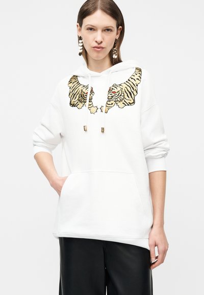 Γυναίκα που φορεί λευκή φούτερ με σχέδια τίγρης στο στήθος, μακριά μανίκια, τσέπη καγκουρό και μαύρο παντελόνι, σε απλό φόντο.