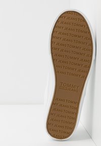 Bijela potplata tenisica s teksturiranom smeđom gumom, s utisnutim uzorkom "TOMMY JEANS" u ponavljanju i čistim, elegantnim dizajnom.