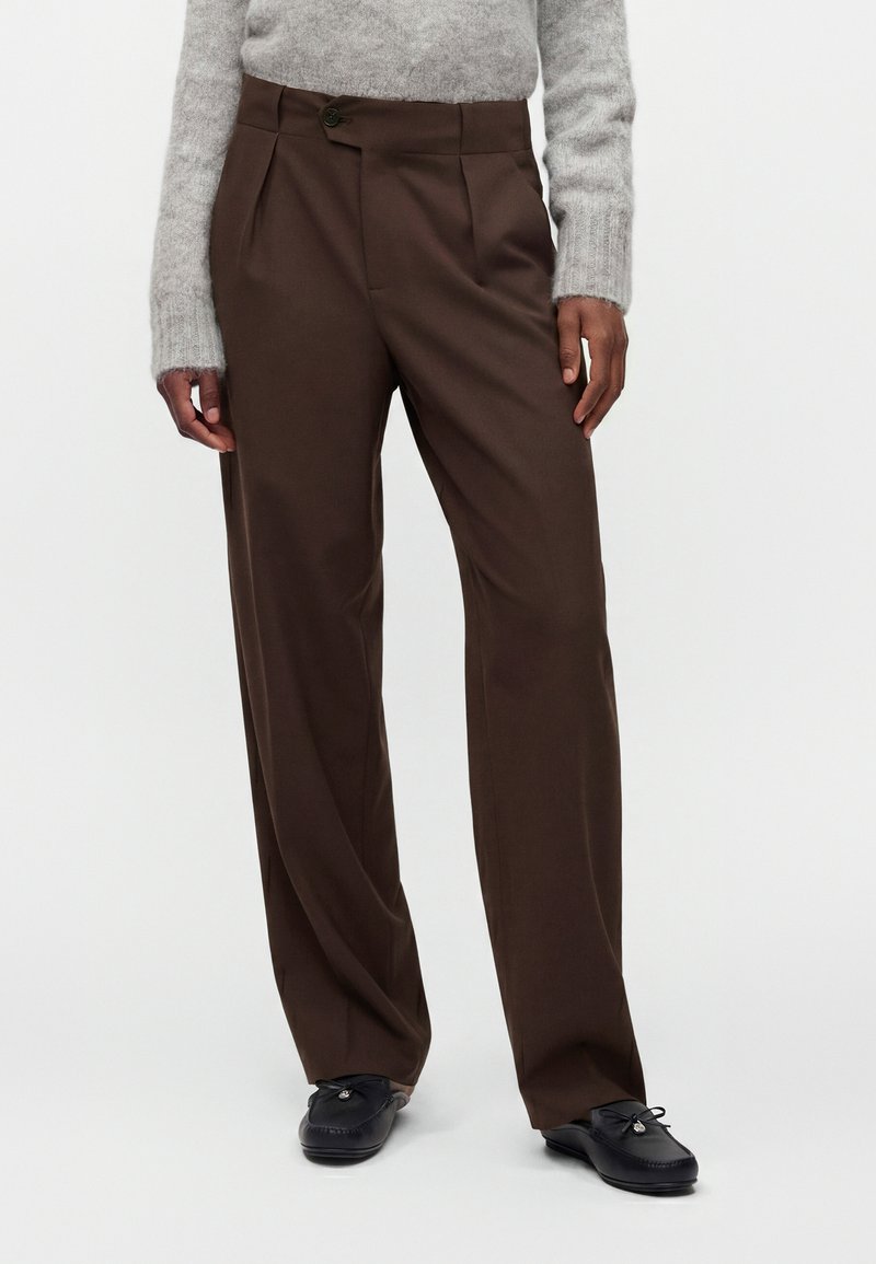 Personne portant un pantalon marron ample, un pull gris clair duveteux et des mocassins noirs, debout devant un fond clair et uni.