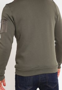 Olive green sweatshirt with a rounded neckline, ribbed cuffs, and hem, featuring a zippered pocket on the left sleeve. Soft, textured fabric.