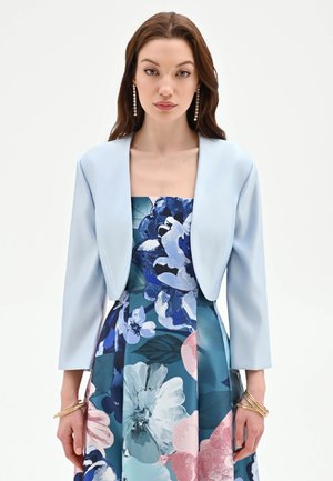 Donna con capelli lunghi castani, orecchini pendenti, blazer corto azzurro pallido e abito senza spalline con grande stampa floreale blu e rosa.