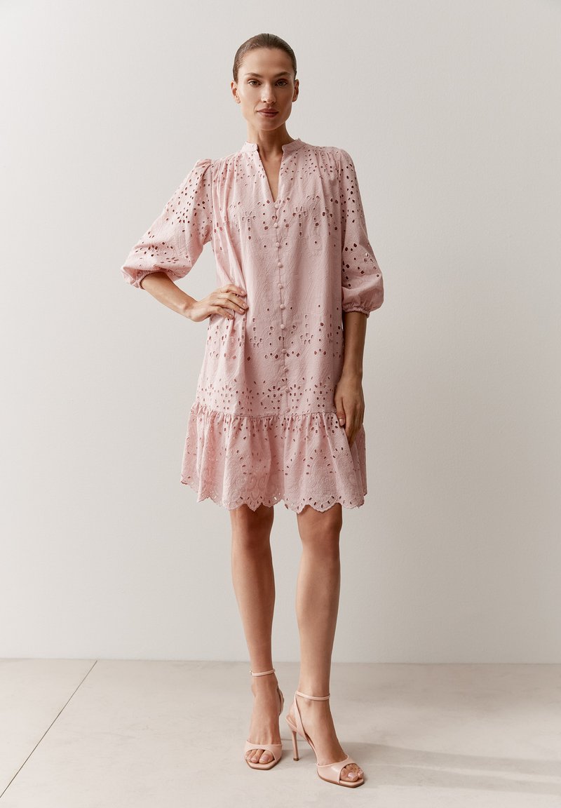 Woman wearing a knee-length pink eyelet dress with puffed sleeves and ruffled hem, paired with strappy nude high-heeled sandals.
