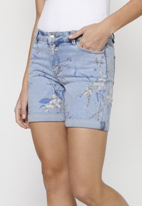 Pantalones cortos de mezclilla azul claro con patrones florales en azul y beige. Con dobladillo enrollado y bolsillos frontales, detalles de botón plateado y cremallera.