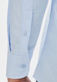 Jasnoniebieska, długa koszula z długim rękawem o tkanej strukturze. Posiada zaokrąglony mankiet z jednym guzikiem oraz lekko zwężający się kształt przy nadgarstku.