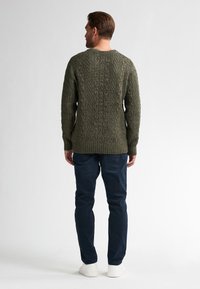 Sweter dziergany w ciemnozielonym kolorze z wzorem w warkocze, o okrągłym dekolcie i ściągaczami. W połączeniu z ciemnoniebieskimi spodniami i białymi butami.