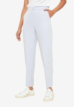 Personne portant un pantalon chevilles sur mesure bleu clair avec poches latérales et des baskets blanches, debout devant un fond uni.