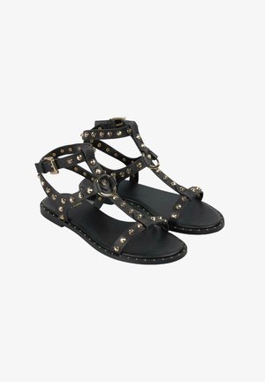 Sandales plates noires avec des accents dorés cloutés, présentant un design à bride T, un talon ouvert et une semelle texturée. Boucle ajustable à la cheville incluse.