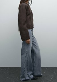 En person klädd i en mörkbrun knäppt jacka och vida ljusblå jeans står på ett mörkt fläckigt golv mot en ljusgrå vägg.