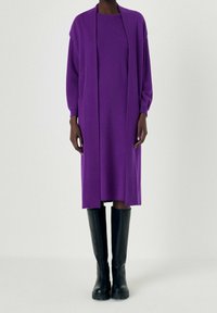 Lungo vestito viola con texture a coste, sovrapposto a un cardigan aperto davanti con maniche larghe e tasche laterali, abbinato a stivali neri fino al ginocchio.