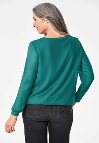 Blusa slanciata color teal con maniche lunghe, scollo rotondo e polsini arricciati. Abbinata a pantaloni in denim scuro con cuciture decorative sulle tasche.