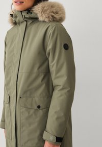 Parka verde oliva con capucha forrada de pelo sintético, múltiples bolsillos y detalles en negro. Presenta un logo en la manga y puños texturizados.