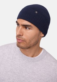 Marine gebreide beanie met gebreide textuur, voorzien van een klein logo-label aan de zijkant; gedragen met een lichtgrijze crewneck trui.