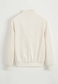 Veste blanche texturée avec un motif chevrons, col montant, poignets côtelés et taille côtelée. Elle possède une poche sur la manche gauche.