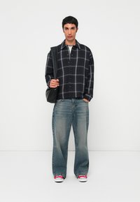 Persona vestida con una camisa de cuadros negra, jeans de pierna ancha y deslavados, zapatillas rojas y llevando un bolso negro de hombro sobre un fondo blanco.