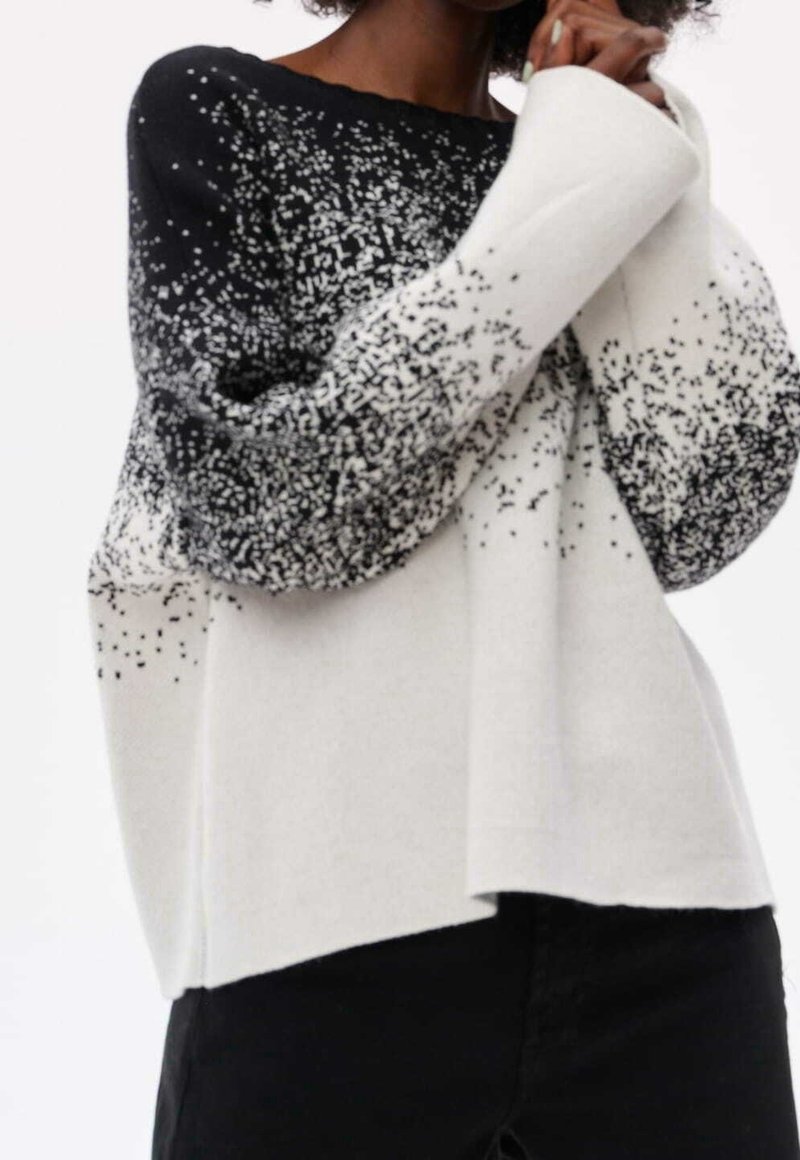 Personne portant un pull en maille noir et blanc tacheté avec des manches longues et un pantalon noir, mains près du visage.
