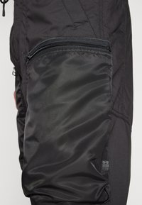 Černé cargo kalhoty s kapsou na zip. Materiál je texturovaný a hladký. Mezi vlastnosti patří elastický manžet a nášivka s logem G-Star.