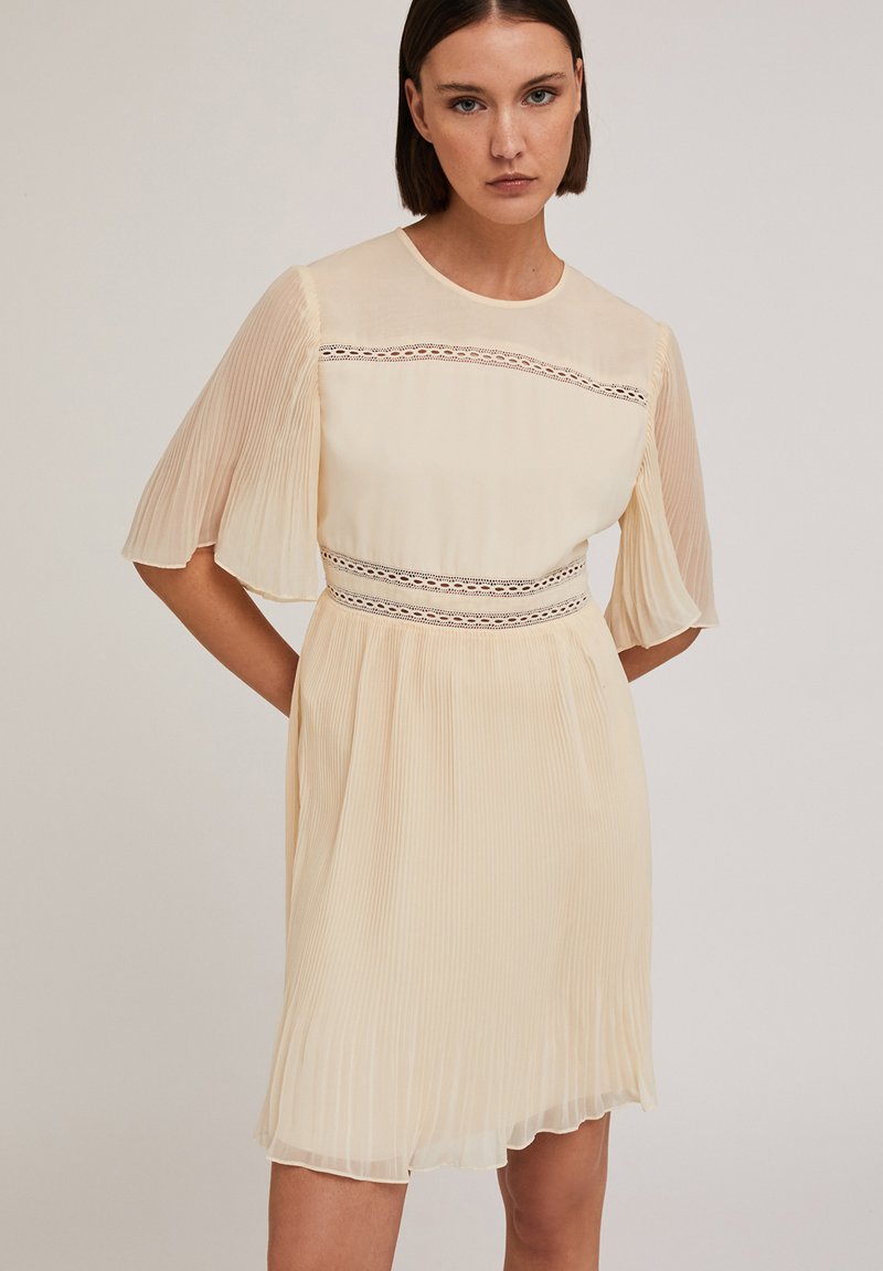 Vestito plissettato color crema chiaro con maniche a farfalla corte e semi-trasparenti. Il bordo intrecciato orizzontale in vita aggiunge un tocco di dettaglio alla silhouette fluida.