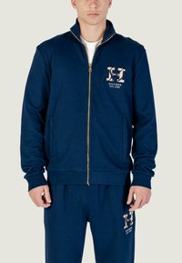 Giacca con zip color navy in tessuto morbido, con colletto alto, tasche laterali e dettaglio logo sul petto. Texture liscia e vestibilità casual.