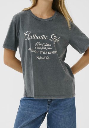Vrouw draagt een vervaagde donkergrijze T-shirt met witte tekst "Authentic Style" en blauwe spijkerbroek, staand met de handen achter haar rug.