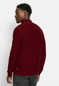 Burgunderfarbener Pullover mit gerippter Struktur, Reißverschlusskragen und seitlichem Streifendetail. Verfügt über eingesetzte Ärmel und einen taillierten Saum.