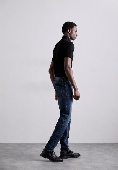Svart polo skjorta, blå jeans och svarta loafers. Slim fit, raka ben jeans, slät textur, subtila sömnadsdetaljer. Neutral bakgrund.