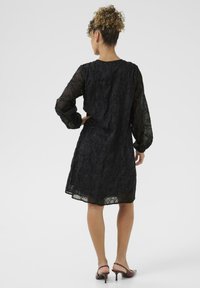Femeie cu păr ondulat, îmbrăcată într-o rochie lungă cu mâneci lungi, dintr-un material transparent, cu modele florale, stând cu o mână pe șold și purtând tocuri vișinii.