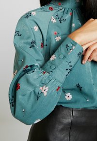 Blusa floreale color turchese con collo alto, maniche a sbuffo e polsini bottonati. Presenta piccoli fiori rossi e bianchi su un tessuto liscio.