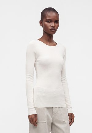Femme aux cheveux courts portant un haut blanc ajusté à manches longues et un pantalon beige clair, se tenant devant un fond blanc uni.