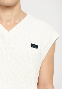 Biały, dziany bezrękawnik z wzorem w romby oraz małą czarną metką marki na klatce piersiowej. Miękka tekstura i luźny krój.