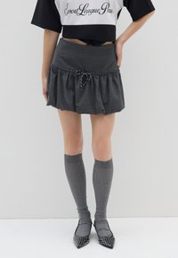 Mini falda de tela gris con un dobladillo volante, que cuenta con una cintura ajustable con cordón. Combinada con calcetas gris de canalé hasta la rodilla y tacones con puntas afiladas y tachuelas.