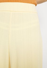 Gros plan d’une personne portant un pantalon côtelé taille haute jaune pâle et un haut côtelé orange sur un fond uni.