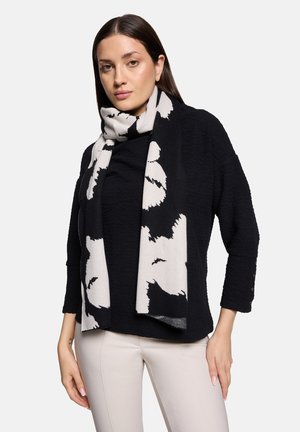 Frau trägt einen schwarzen, strukturierten Langarmpullover, beige Hose und einen schwarz-weiß gemusterten abstrakten Schal, der um ihren Hals gewickelt ist.