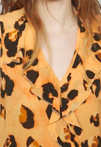 Orange blouse with black and brown leopard print, featuring ruffled detailing along the neckline and a V-shaped cut. Smooth texture.