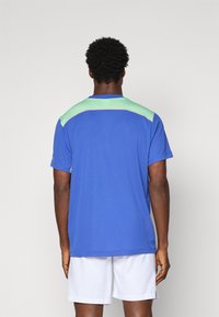 Man wearing blue and green sports shirt and white shorts, facing away against plain light gray background.